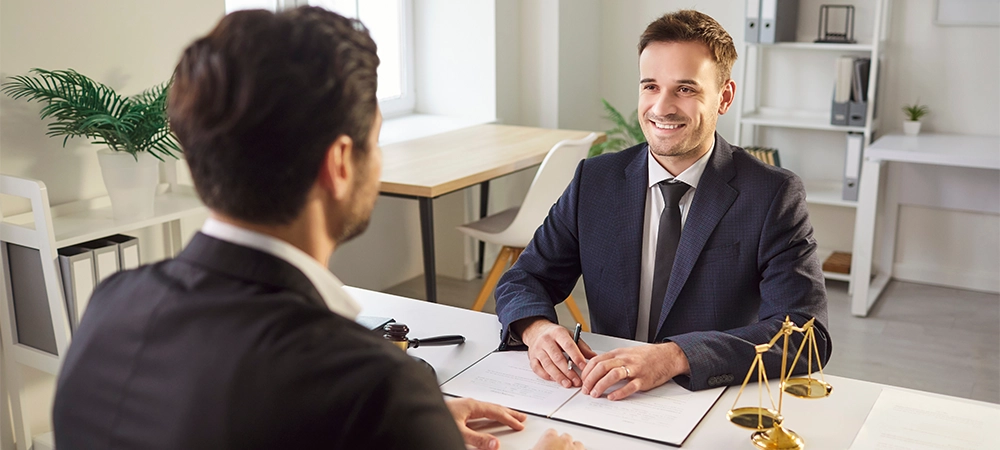 Smiling male lawyer in suit sitting at desk with legal documents, consulting client in modern office. Professional attorney listening attentively, providing advice and legal assistance during meeting.