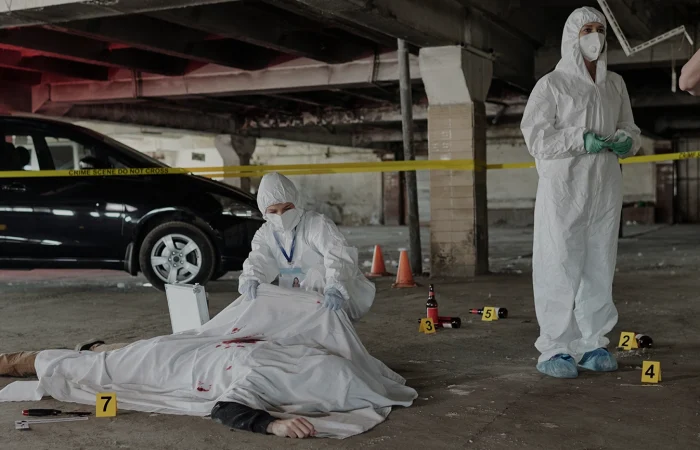 Young policeman communicating with forensic expert at crime scene with dead body covered with white sheet in parking area