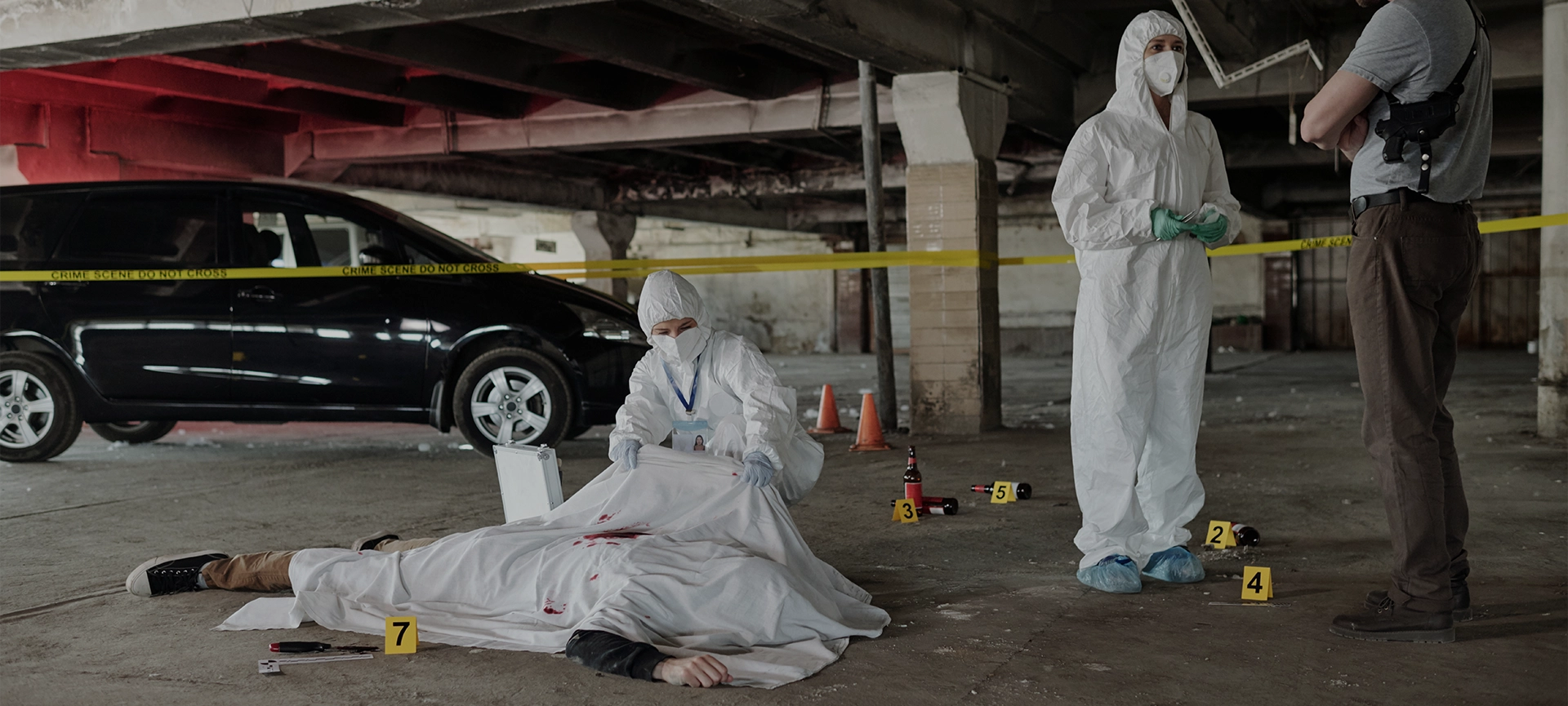Young policeman communicating with forensic expert at crime scene with dead body covered with white sheet in parking area