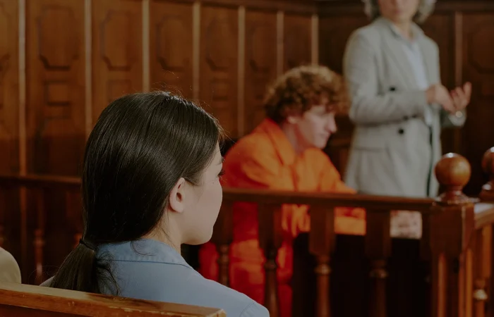 Caucasian teenage boy with curly hair sitting in defendant box wearing orange jumpsuit, middle aged Caucasian woman standing and gesturing as lawyer, diverse adults seated in courtroom observing proceedings