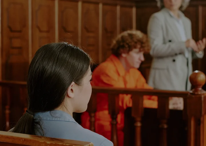 Caucasian teenage boy with curly hair sitting in defendant box wearing orange jumpsuit, middle aged Caucasian woman standing and gesturing as lawyer, diverse adults seated in courtroom observing proceedings