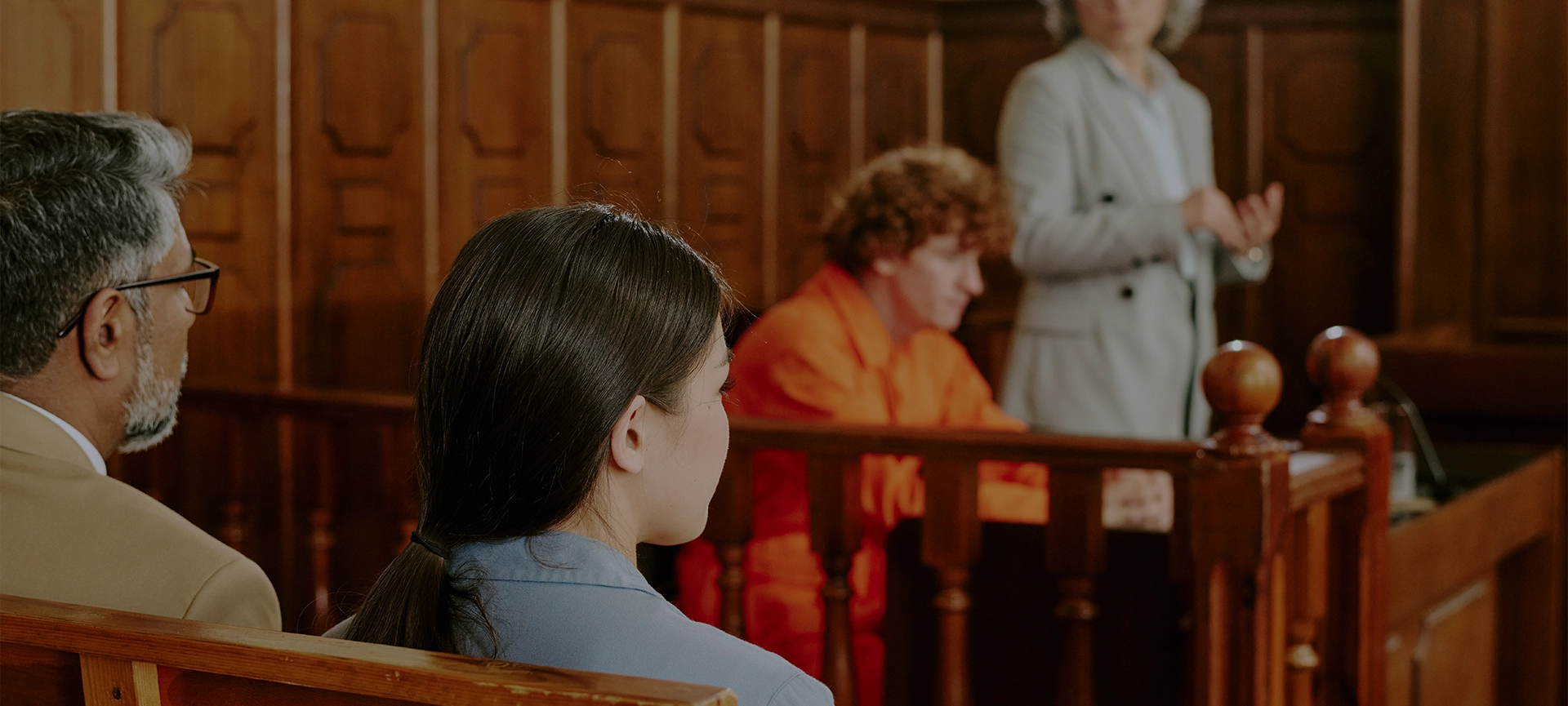 Caucasian teenage boy with curly hair sitting in defendant box wearing orange jumpsuit, middle aged Caucasian woman standing and gesturing as lawyer, diverse adults seated in courtroom observing proceedings