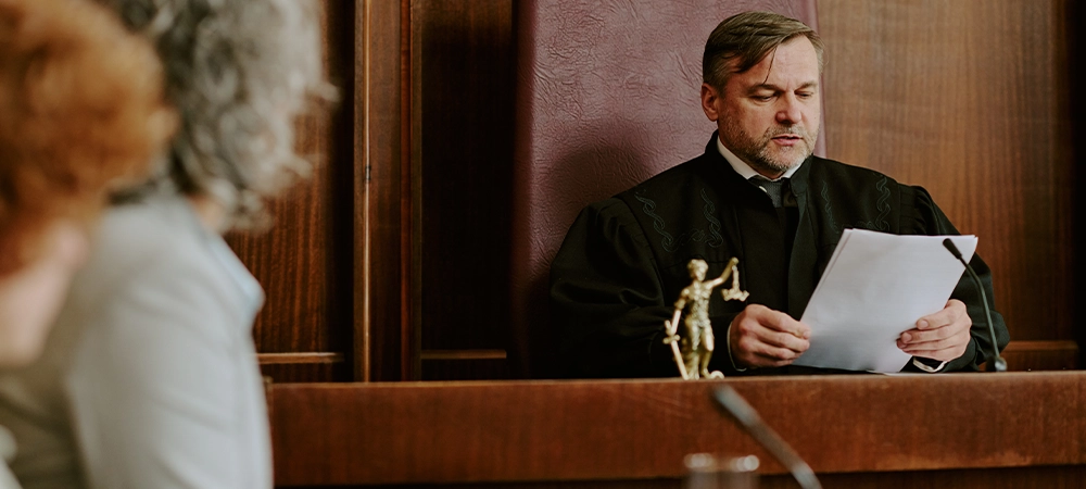 Caucasian middle aged man judge reading legal documents in courtroom while two women listening in foreground, legal proceedings taking place in judicial setting with focus on authority