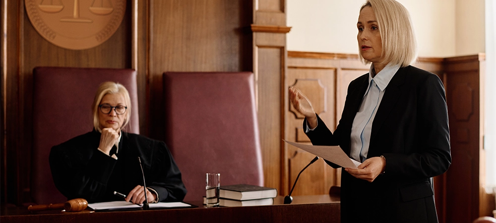 Confident blond female prosecutor with juridical document looking at suspect and his attorney while standing against impartial judge