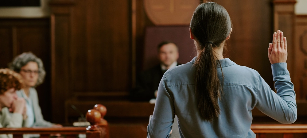 Caucasian young adult woman standing in courtroom raising right hand while swearing oath during legal proceedings with judge and attorneys observing in background