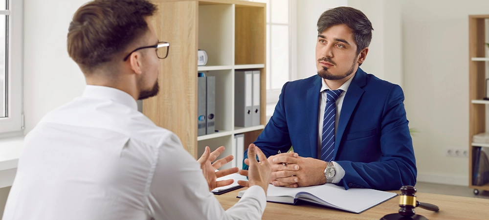 Young man talking to a professional lawyer. Serious attorney in a formal suit sitting at his office desk and listening to a client telling about his problem. Law consultation, legal advice concept