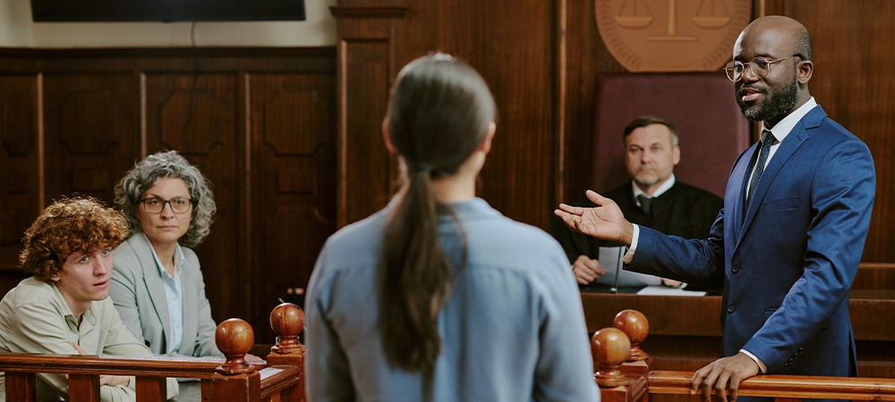 Black middle aged man in suit addressing Caucasian young adult woman standing in witness box, while Caucasian middle aged woman and Caucasian young adult man observing in courtroom