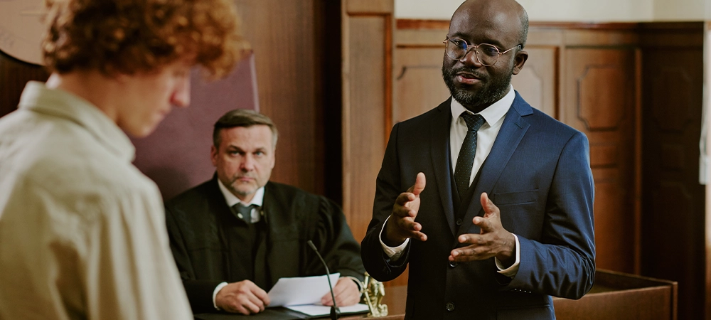 Black middle aged man in suit gesturing while speaking in courtroom, Caucasian teenage boy standing in foreground, Caucasian middle aged male judge sitting behind bench observing